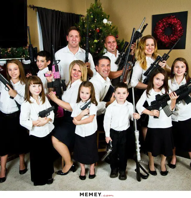 Five adults and seven children, formally dressed in black pants or skirts and white shirts and blouses, each proudly displaying a gun, rifle, or automatic weapon posed in front of a Christmas tree. 