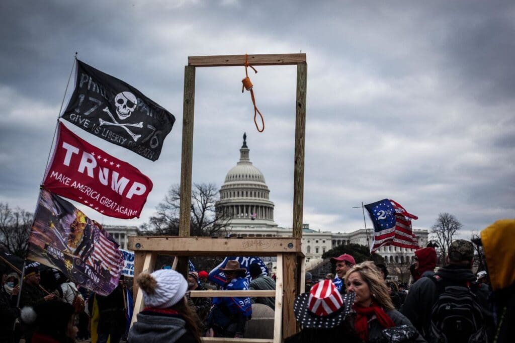 The Capitol Building dome and the gallows with a rope hanging from it, along with a Trump flag and skull and crossbones, with the angry mob on January 6, 2021.